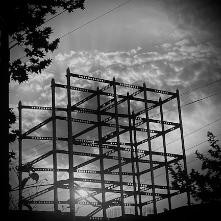 A silhouetted steel framework of a building under construction is set against a dramatic sky filled with textured clouds. Sunlight peeks through the gaps, creating a stark contrast with the dark structure. Silhouetted leaves and branches from nearby trees border the scene.