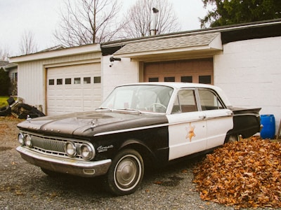 A vintage black and white patrol car is parked in front of a garage. Leaves are piled up near the vehicle, and there is a blue barrel to the side. The garage has a white exterior and a closed door with small windows. Bare trees and scattered leaves suggest a late autumn setting.