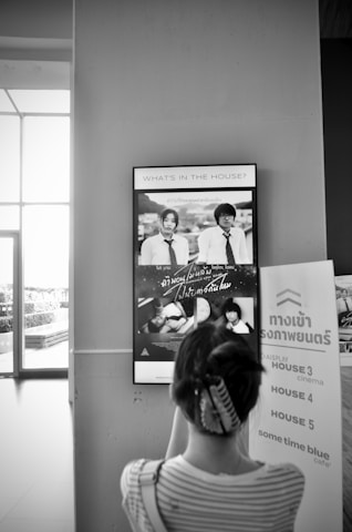 A person is looking at a display advertisement mounted on a wall. The advertisement features two individuals wearing ties and white shirts. Below the main image, there are smaller images, including one of the same individuals in different scenes. There are also signs next to the person, indicating directions to cinema houses and a cafe.
