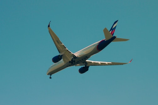 An airplane is flying overhead against a clear blue sky. The aircraft has a sleek design with a prominent tail fin displaying a color pattern. The engines are visible under the wings, and the plane is angled in a way that reveals the word 'SLOT' on its underbelly.