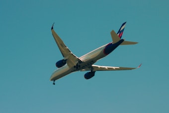 An airplane is flying overhead against a clear blue sky. The aircraft has a sleek design with a prominent tail fin displaying a color pattern. The engines are visible under the wings, and the plane is angled in a way that reveals the word 'SLOT' on its underbelly.