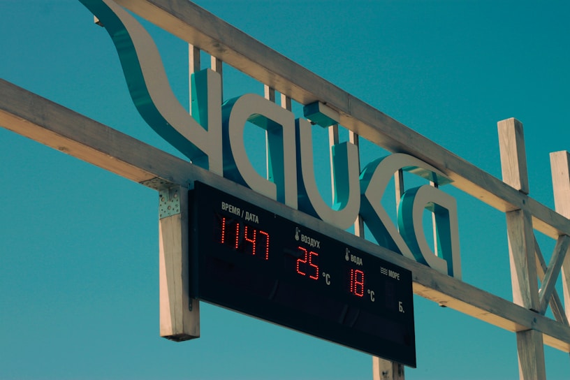A close-up view of a digital sign with red LED numerals displaying time and temperature, mounted on a wooden frame against a clear blue sky. The sign shows the time as 11:47 and the air and water temperatures as 25°C and 18°C, respectively. Additionally, there is some text in another language, likely related to the display information.
