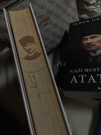 A vertical book with a wooden cover featuring an engraved portrait and signature. Beside it lies a book with a partially visible cover showing a man's face. There is a patterned bag in the background.