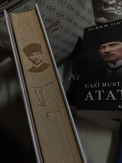 A vertical book with a wooden cover featuring an engraved portrait and signature. Beside it lies a book with a partially visible cover showing a man's face. There is a patterned bag in the background.