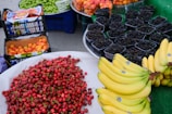 A vibrant display of various fruits including bananas, red cherries, peaches, green grapes, and mulberries arranged on market tables. The cherries are piled high in a large mound, bananas are neatly stacked in clusters, and the mulberries are individually packaged in small containers.