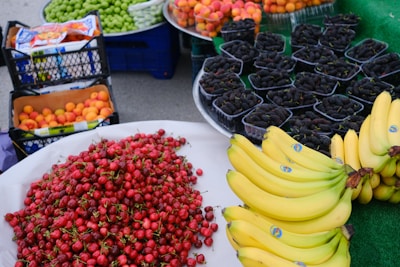A vibrant display of various fruits including bananas, red cherries, peaches, green grapes, and mulberries arranged on market tables. The cherries are piled high in a large mound, bananas are neatly stacked in clusters, and the mulberries are individually packaged in small containers.