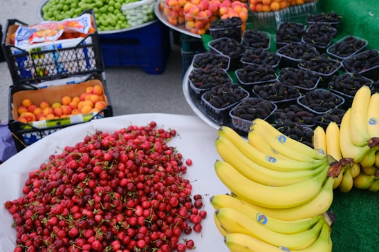 A vibrant display of various fruits including bananas, red cherries, peaches, green grapes, and mulberries arranged on market tables. The cherries are piled high in a large mound, bananas are neatly stacked in clusters, and the mulberries are individually packaged in small containers.