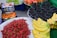 A vibrant display of various fruits including bananas, red cherries, peaches, green grapes, and mulberries arranged on market tables. The cherries are piled high in a large mound, bananas are neatly stacked in clusters, and the mulberries are individually packaged in small containers.