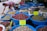A market stall features an array of large blue plastic bowls filled with various dried herbs and flowers. The bowls are filled to the brim with items like pink rose buds, green leaves, and lemon balm, as indicated by handwritten yellow signs. A person in a pink shirt is reaching toward some items outside the main collection of bowls. Behind the bowls, there are additional bins and sacks filled with similar dried goods.