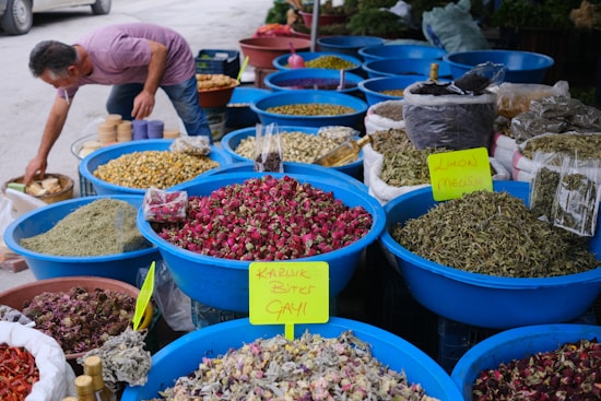 A market stall features an array of large blue plastic bowls filled with various dried herbs and flowers. The bowls are filled to the brim with items like pink rose buds, green leaves, and lemon balm, as indicated by handwritten yellow signs. A person in a pink shirt is reaching toward some items outside the main collection of bowls. Behind the bowls, there are additional bins and sacks filled with similar dried goods.