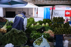A market stall with an assortment of fresh green vegetables, including artichokes, lettuce, herbs, and onions. A vendor is seen from behind wearing a dark jacket with yellow lettering. The stall is shaded by an umbrella, and there are other market signs and stalls in the background.