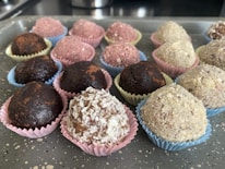 An assortment of round, chocolate-colored treats placed in colorful, decorative paper cups on a speckled gray tray. The sweets vary in texture, some dusted with cocoa or covered in shredded coconut and other ingredients.