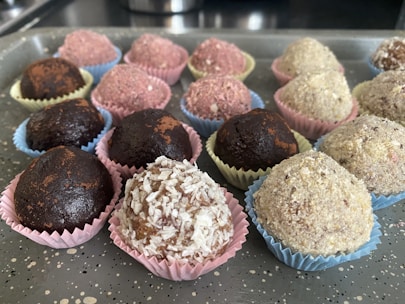 An assortment of round, chocolate-colored treats placed in colorful, decorative paper cups on a speckled gray tray. The sweets vary in texture, some dusted with cocoa or covered in shredded coconut and other ingredients.