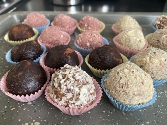 An assortment of round, chocolate-colored treats placed in colorful, decorative paper cups on a speckled gray tray. The sweets vary in texture, some dusted with cocoa or covered in shredded coconut and other ingredients.
