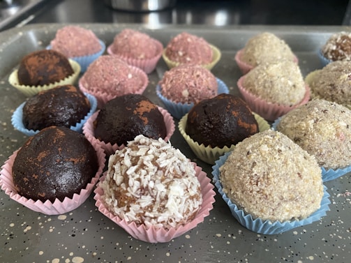 An assortment of round, chocolate-colored treats placed in colorful, decorative paper cups on a speckled gray tray. The sweets vary in texture, some dusted with cocoa or covered in shredded coconut and other ingredients.