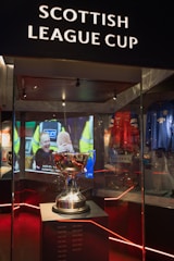 A trophy is displayed in a glass case under the sign 'Scottish League Cup'. Reflections and ambient lighting create a dramatic atmosphere. In the background, there is a video screen showing a sports-related scene. Various sports memorabilia, including jerseys, surround the main display.
