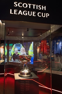 A trophy is displayed in a glass case under the sign 'Scottish League Cup'. Reflections and ambient lighting create a dramatic atmosphere. In the background, there is a video screen showing a sports-related scene. Various sports memorabilia, including jerseys, surround the main display.