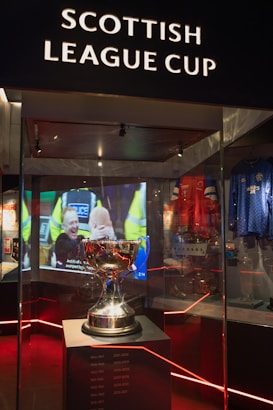 A trophy is displayed in a glass case under the sign 'Scottish League Cup'. Reflections and ambient lighting create a dramatic atmosphere. In the background, there is a video screen showing a sports-related scene. Various sports memorabilia, including jerseys, surround the main display.