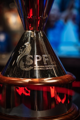 A polished trophy with the inscription 'SPFL Scottish Professional Football League' engraved on it. The trophy features a silver surface with reflective qualities and a decorative base with red accents. The backdrop includes blurred, colorful lights and shapes.
