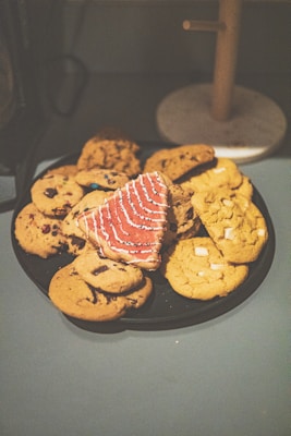 A variety of cookies are arranged on a dark platter. The assortment includes chocolate chip cookies, cookies with white chocolate chunks, and a distinct pink cookie shaped like a tree with white icing decoration.