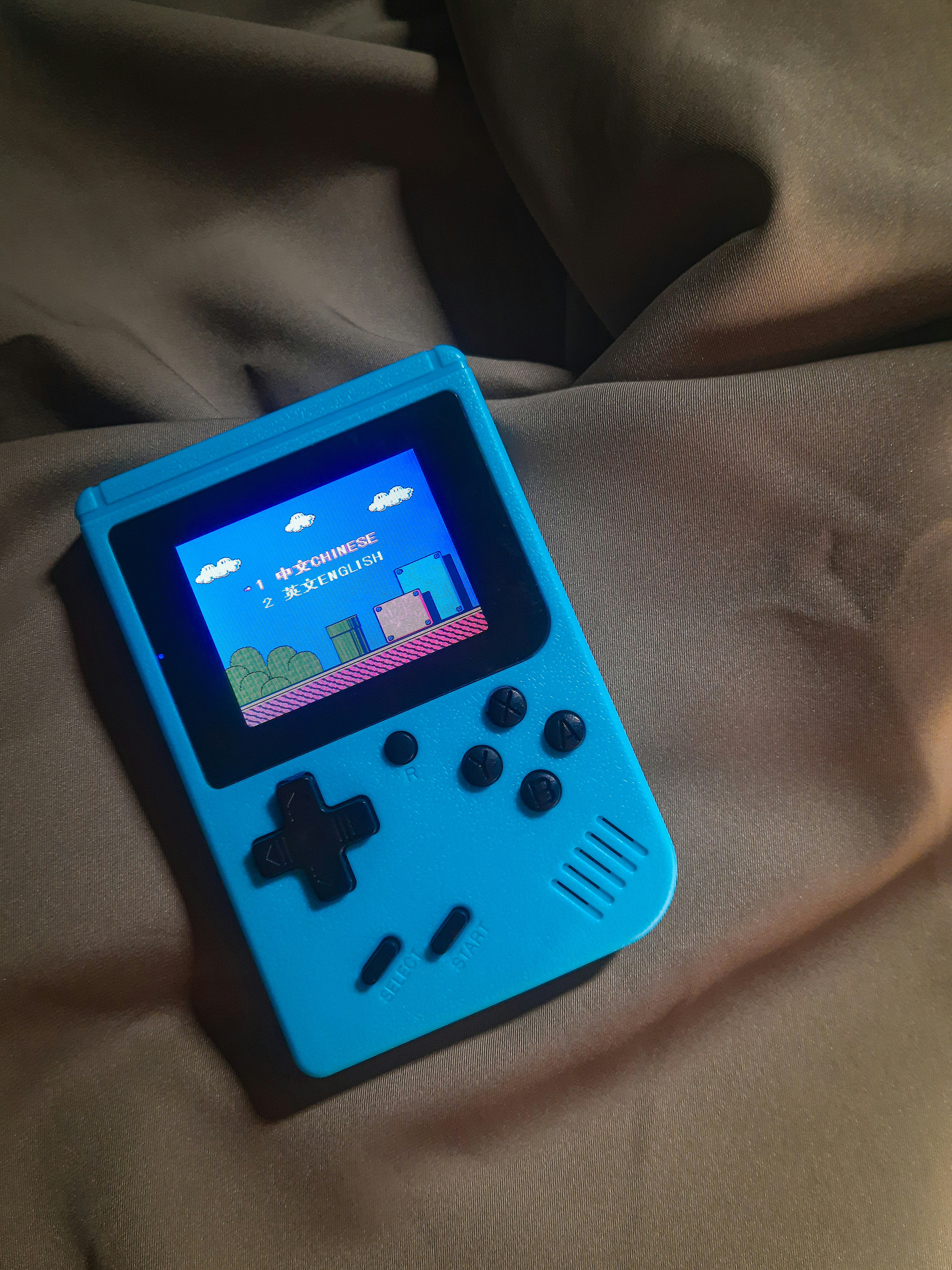 A handheld gaming device with a bright blue casing rests on a soft, dark fabric background. The screen displays a pixelated video game scene with options to select either Chinese or English language. The layout includes a classic D-pad and buttons, reminiscent of retro gaming consoles.