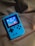 A handheld gaming device with a bright blue casing rests on a soft, dark fabric background. The screen displays a pixelated video game scene with options to select either Chinese or English language. The layout includes a classic D-pad and buttons, reminiscent of retro gaming consoles.