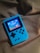 A handheld gaming device with a bright blue casing rests on a soft, dark fabric background. The screen displays a pixelated video game scene with options to select either Chinese or English language. The layout includes a classic D-pad and buttons, reminiscent of retro gaming consoles.