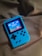 A handheld gaming device with a bright blue casing rests on a soft, dark fabric background. The screen displays a pixelated video game scene with options to select either Chinese or English language. The layout includes a classic D-pad and buttons, reminiscent of retro gaming consoles.