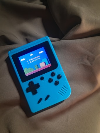 A handheld gaming device with a bright blue casing rests on a soft, dark fabric background. The screen displays a pixelated video game scene with options to select either Chinese or English language. The layout includes a classic D-pad and buttons, reminiscent of retro gaming consoles.
