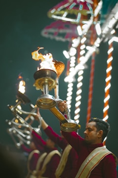 A man in traditional attire holds a burning lamp as part of a ceremonial ritual. In the background, several other individuals are participating in the same activity, with a colorful, illuminated structure providing a vibrant backdrop.