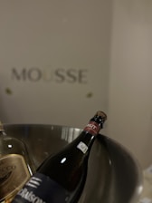 Two bottles of sparkling wine are nestled in a metallic ice bucket. The label on one bottle is marked as Brut. The background is minimal, featuring the text 'Mousse' on the wall.