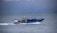 A group of people wearing life jackets and protective clothing are seated in a fast-moving speedboat on a large body of water. The boat has a flag with a blue cross emblem. Behind the boat, there are misty mountains partially covered with greenery and patches of snow.