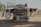 A small, open-fronted roadside shop or kiosk displaying various crafts and goods. The structure is adorned with colorful cloths and tapestries depicting camels and desert scenes. An Egyptian flag is prominently displayed above the shop. Several small statues and trinkets are arranged neatly on shelves. A plastic chair is placed to the left, and various items hang from the awning and walls.
