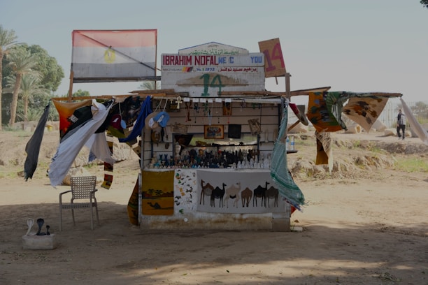 A small, open-fronted roadside shop or kiosk displaying various crafts and goods. The structure is adorned with colorful cloths and tapestries depicting camels and desert scenes. An Egyptian flag is prominently displayed above the shop. Several small statues and trinkets are arranged neatly on shelves. A plastic chair is placed to the left, and various items hang from the awning and walls.