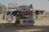 A small, open-fronted roadside shop or kiosk displaying various crafts and goods. The structure is adorned with colorful cloths and tapestries depicting camels and desert scenes. An Egyptian flag is prominently displayed above the shop. Several small statues and trinkets are arranged neatly on shelves. A plastic chair is placed to the left, and various items hang from the awning and walls.