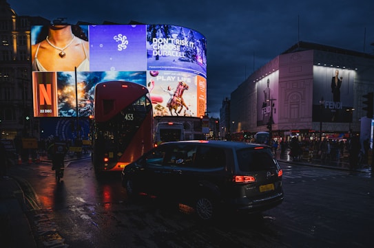 A busy city street at night featuring bright advertisements on large digital billboards. The advertising signs include a mix of images and text promoting various products such as a necklace, a Netflix release, Duracell batteries, and a horse-themed advertisement. The street is crowded with vehicles including a red double-decker bus and multiple cars.