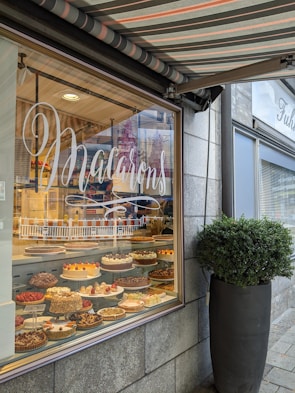 A bakery window display with a variety of ornate cakes and pastries arranged inside. The glass window features the word 'Macarons' in elegant script, and there is a large potted plant next to the window on the stone sidewalk. A striped awning shades the top of the window, adding a cozy and inviting feel to the shop exterior.