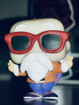 A stylized, cartoonish figure with a large white beard and red sunglasses. The figure is wearing an orange jacket and blue pants, standing upright. The background is blurred, emphasizing the figure.