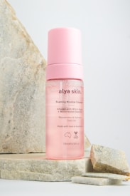 A pink bottle of Alya Skin Foaming Micellar Cleanser is prominently displayed against a light-colored stone backdrop. The bottle has a pink cap and transparent body, showing the liquid inside. The label mentions ingredients like Witch Hazel and Watermelon Seed Oil, and highlights its rejuvenating and hydrating properties. The product is marked as being made in Australia.