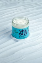 A jar of hair mask with a creamy white substance is displayed on a textured surface. The packaging features a blue label with the brand name 'VITA COCO' and illustrated green leaves.
