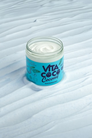 A jar of hair mask with a creamy white substance is displayed on a textured surface. The packaging features a blue label with the brand name 'VITA COCO' and illustrated green leaves.