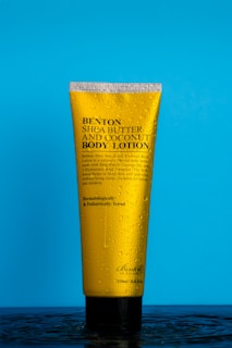 A yellow bottle of Benton Shea Butter and Coconut Body Lotion is standing upright on a bright blue background. The bottle is covered in water droplets, giving it a fresh and clean appearance. The text on the bottle highlights its key ingredients and dermatological testing.