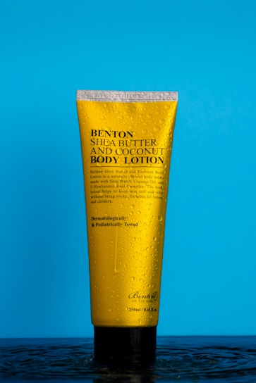 A yellow bottle of Benton Shea Butter and Coconut Body Lotion is standing upright on a bright blue background. The bottle is covered in water droplets, giving it a fresh and clean appearance. The text on the bottle highlights its key ingredients and dermatological testing.