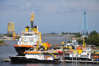 Several boats are docked at a busy industrial harbor. The largest vessel, with a red and black hull and a yellow crane, is prominently positioned alongside smaller boats. In the background, cranes and various industrial structures are visible, with the horizon showcasing buildings and trees.