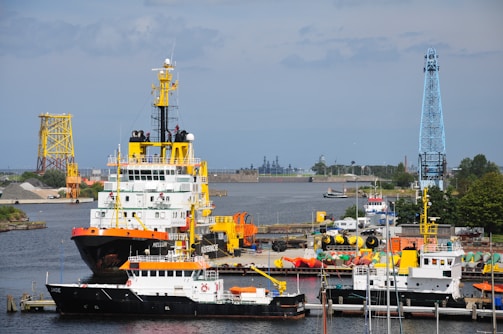 Several boats are docked at a busy industrial harbor. The largest vessel, with a red and black hull and a yellow crane, is prominently positioned alongside smaller boats. In the background, cranes and various industrial structures are visible, with the horizon showcasing buildings and trees.