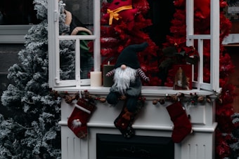 A festive holiday display with a small gnome-like figure sitting on a white mantel decorated with pine cones and stockings. To the left, there is a frosted artificial Christmas tree, while a bright red tree is on the right. An open window frame is present, along with a burning candle and a small framed portrait nearby. The scene conveys a cozy, wintery atmosphere.