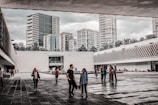An open courtyard with people walking and socializing, surrounded by modern concrete and glass architecture. Numerous high-rise buildings can be seen in the background, and the sky appears overcast. The area is interspersed with trees and landscaping, adding a touch of green.