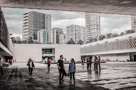 An open courtyard with people walking and socializing, surrounded by modern concrete and glass architecture. Numerous high-rise buildings can be seen in the background, and the sky appears overcast. The area is interspersed with trees and landscaping, adding a touch of green.