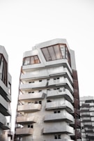 A modern, multi-story apartment building featuring a unique architectural design with geometric shapes and lines. The building has multiple balconies and large windows. Adjacent buildings of a similar architectural style can be seen in the background. The facade has a mix of white and grey tones with wood paneling around the windows.
