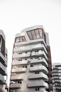 A modern, multi-story apartment building featuring a unique architectural design with geometric shapes and lines. The building has multiple balconies and large windows. Adjacent buildings of a similar architectural style can be seen in the background. The facade has a mix of white and grey tones with wood paneling around the windows.